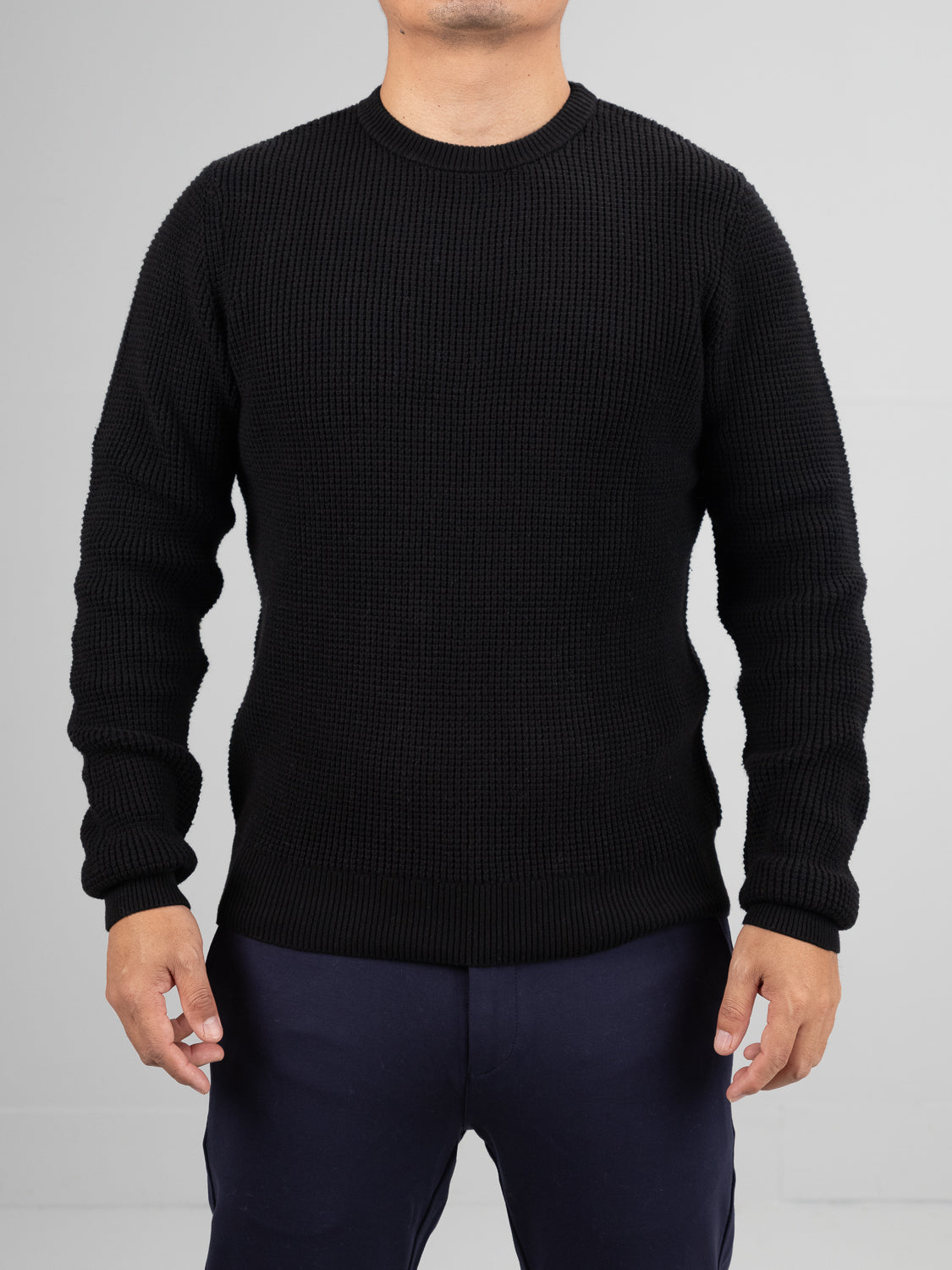 A person wears a black Ragnar Waffle Knit Sweater from ID Menswear, made of 100% cotton, with dark pants against a plain gray background. The sweaters texture contrasts as their hands rest by their sides. The head is not visible in the image.