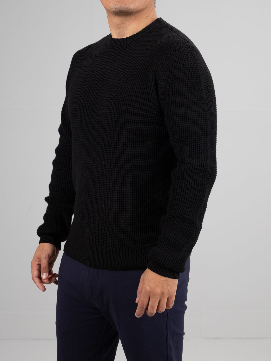 A person in a Ragnar Waffle Knit Sweater by ID Menswear, crafted from 100% cotton, and dark pants stands against a gray background. Only the upper body is visible as the head is slightly turned, obscuring the face.