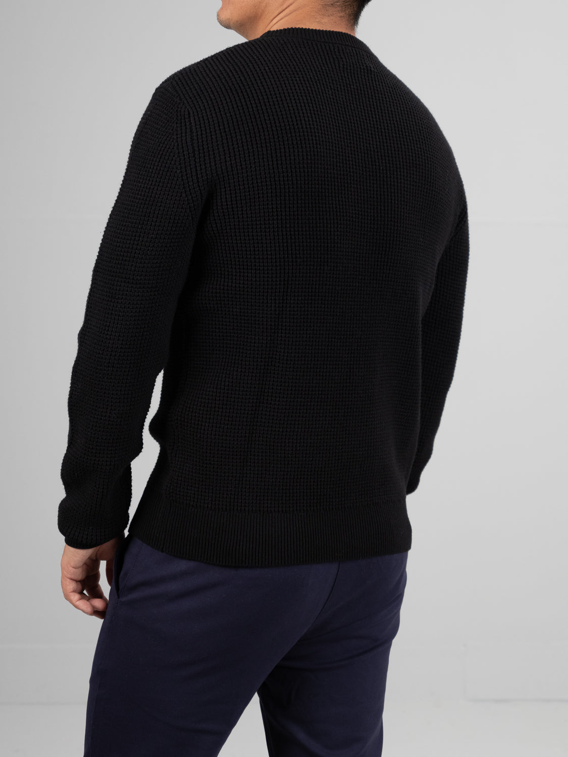 A person stands facing away in a black Ragnar Waffle Knit Sweater, branded by ID Menswear and crafted from 100% cotton with a textured ribbed design, paired with navy pants. The background is plain light gray.