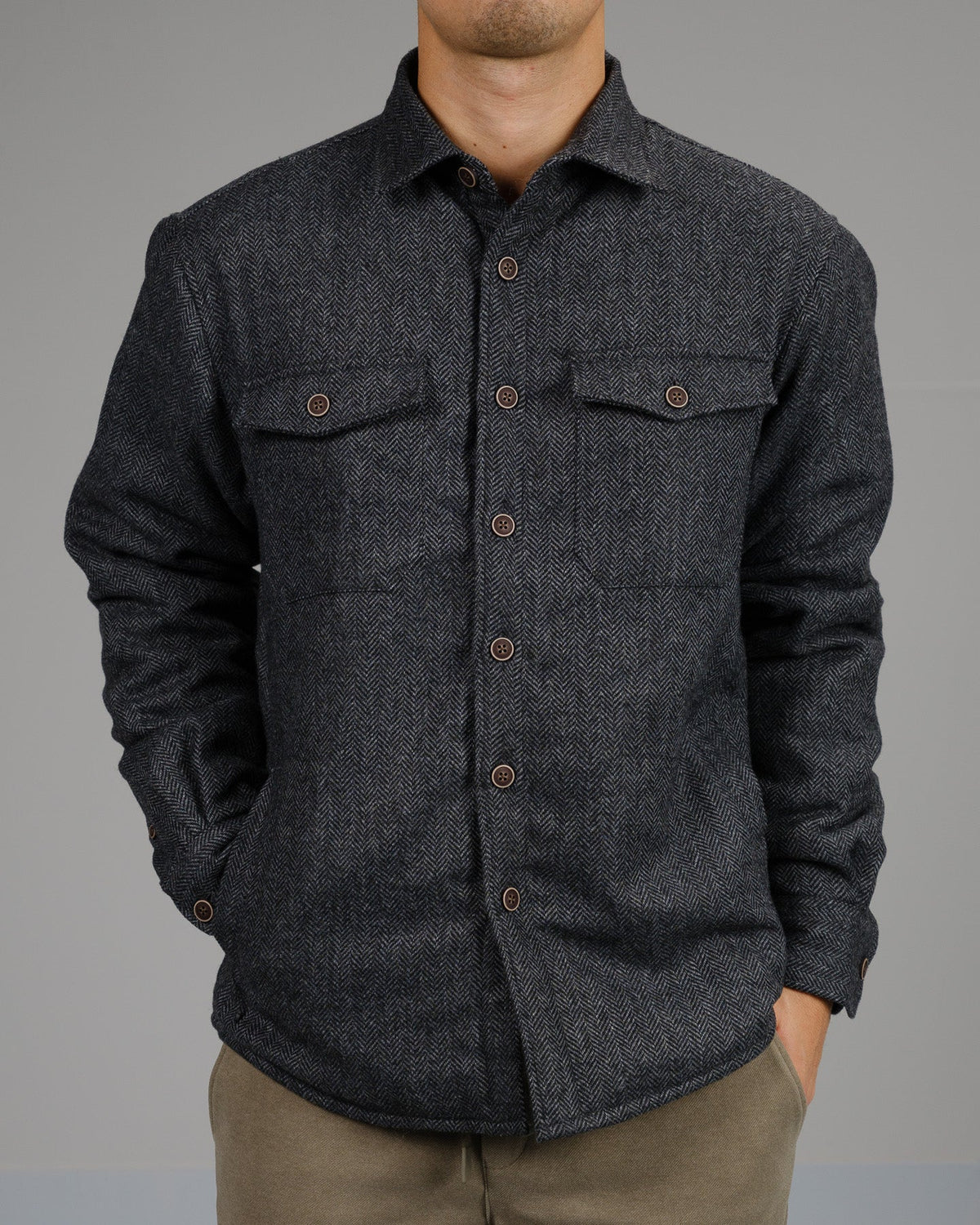 A person wearing a dark gray button-up shirt and light brown pants stands with hands in pockets, layered under the ID Menswear Stuntman Wool Blend Tweed Jacket, against a plain gray background.