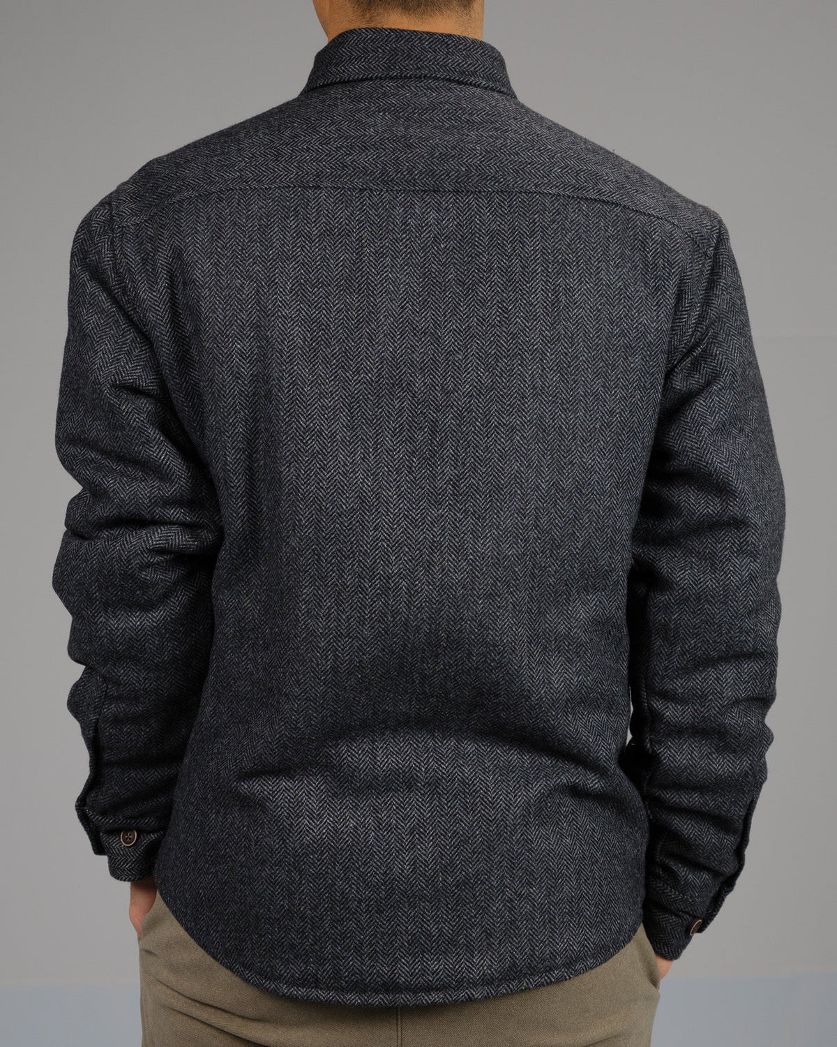 A person is seen from behind wearing the ID Menswear Stuntman Wool Blend Tweed Jacket in dark gray with long sleeves, paired with light brown pants, against a plain gray background.