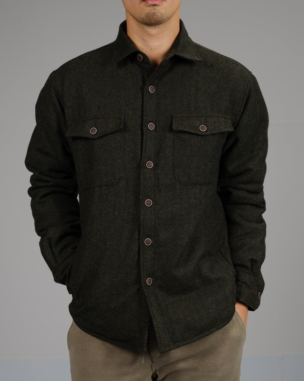 A person wears the ID Menswear Stuntman Wool Blend Tweed Jacket in dark green, featuring two chest pockets and brown buttons, with hands in their light brown pants pockets, standing out against a plain gray background.