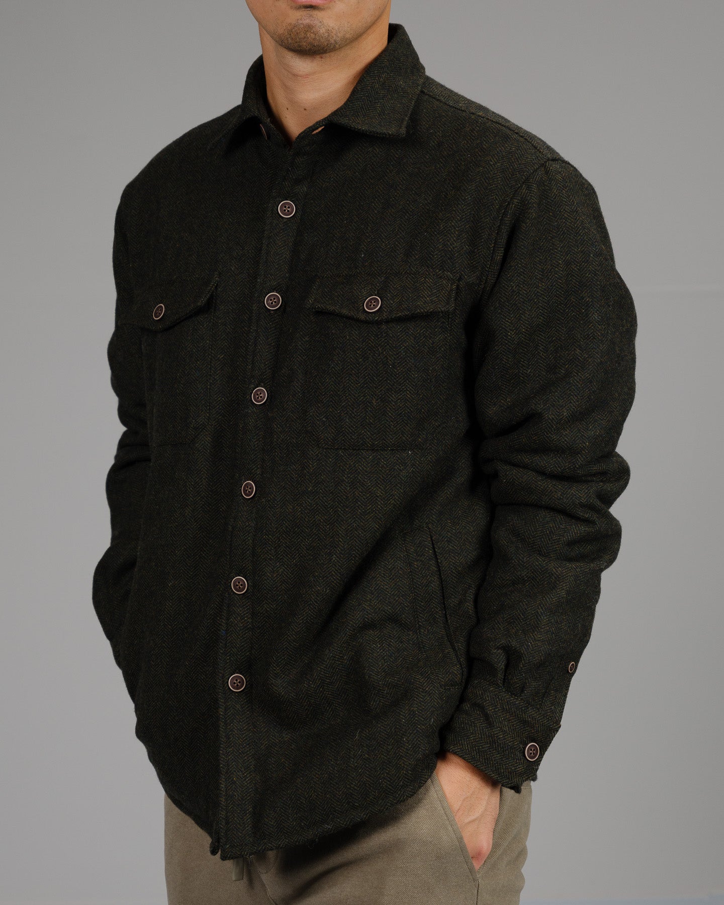 A man in the ID Menswear Stuntman Wool Blend Tweed Jacket with flap chest pockets and beige pants stands against a gray background, his left hand in his pocket and face partly visible, exemplifying timeless outerwear style.