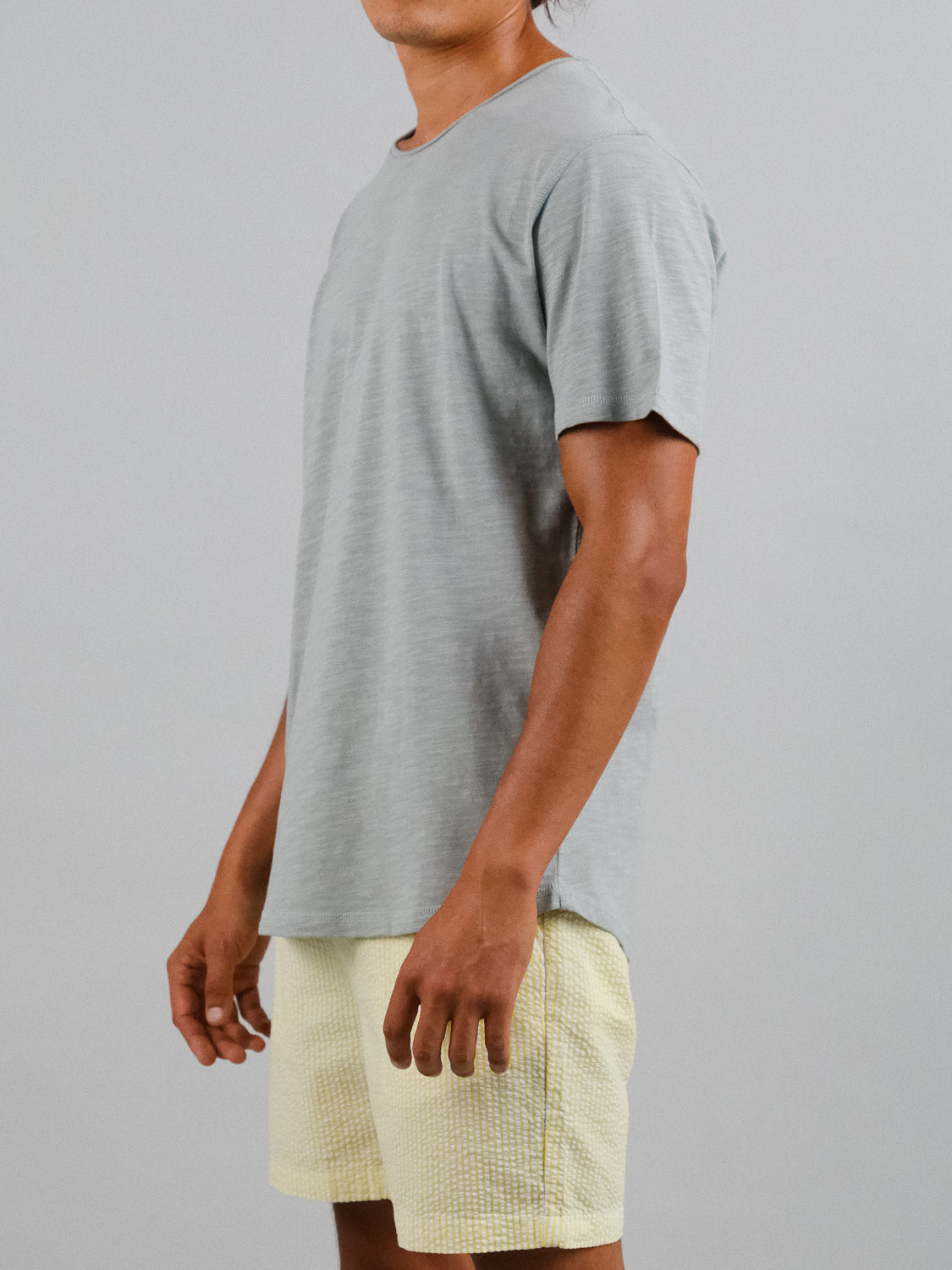 A person in a gray Da Nang Rolled Neck Slub Tee from ID Menswear and light yellow shorts stands against a plain light gray background, slightly facing left with hands relaxed at their sides.