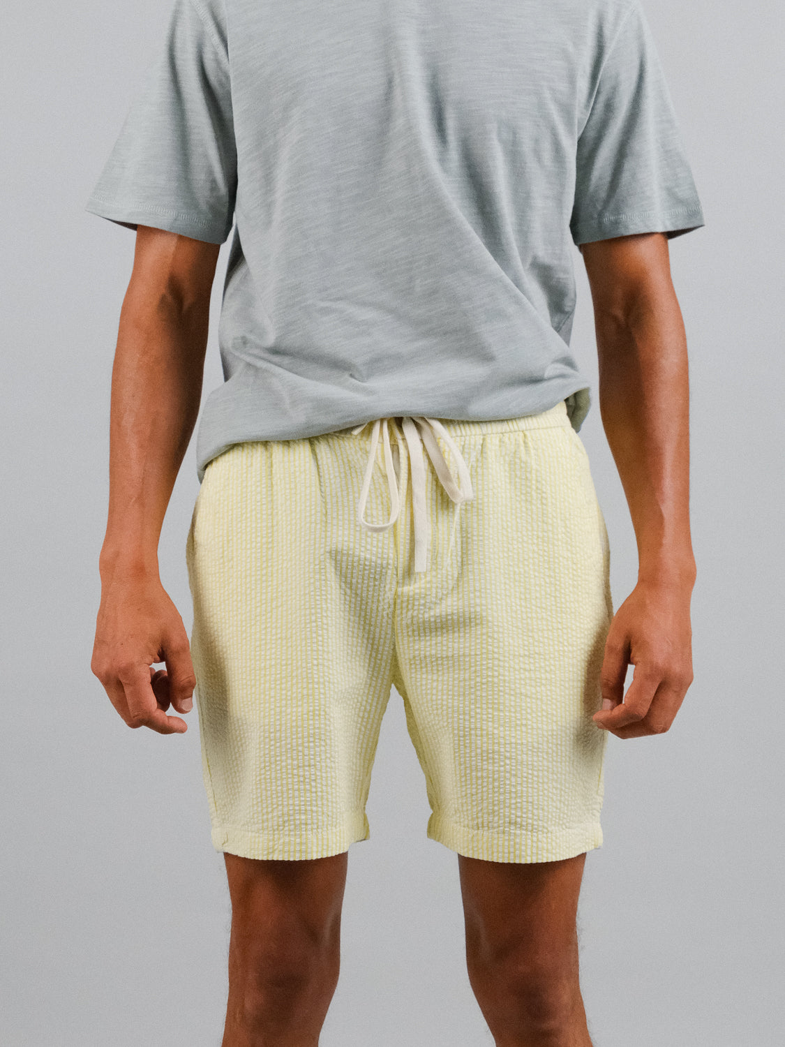 A person displays a warm weather outfit featuring the Ibiza Seersucker Drawstring Short by ID Menswear and a light gray t-shirt against a simple, light gray backdrop. The photo is taken from the neck down.