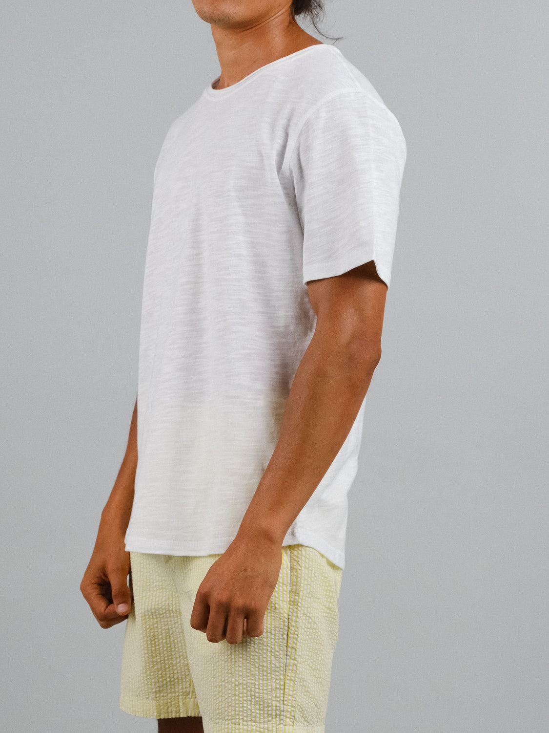 A person models a Da Nang Rolled Neck Slub Tee by ID Menswear with light yellow shorts against a gray backdrop, epitomizing effortless style.