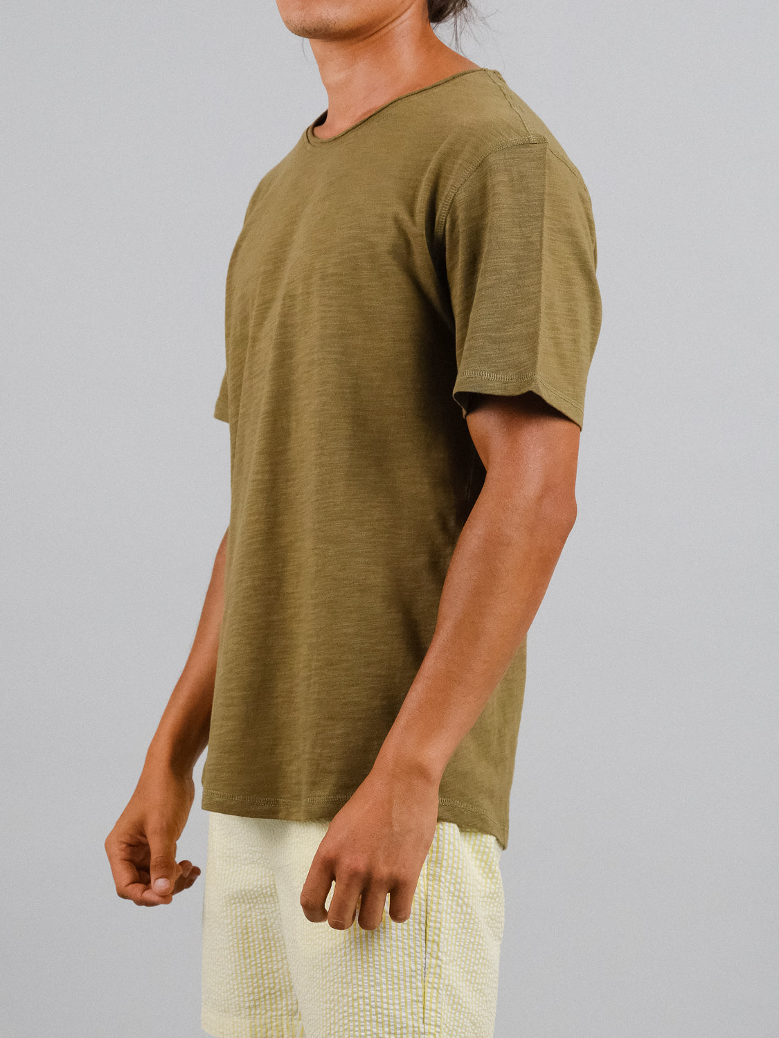 A person wears an olive ID Menswear Da Nang Rolled Neck Slub Tee and pale yellow shorts, standing against a neutral background. The slub cotton fabric accentuates the casual vibe as they relax their arms at the sides. The face is not visible and focus stays on the upper body.