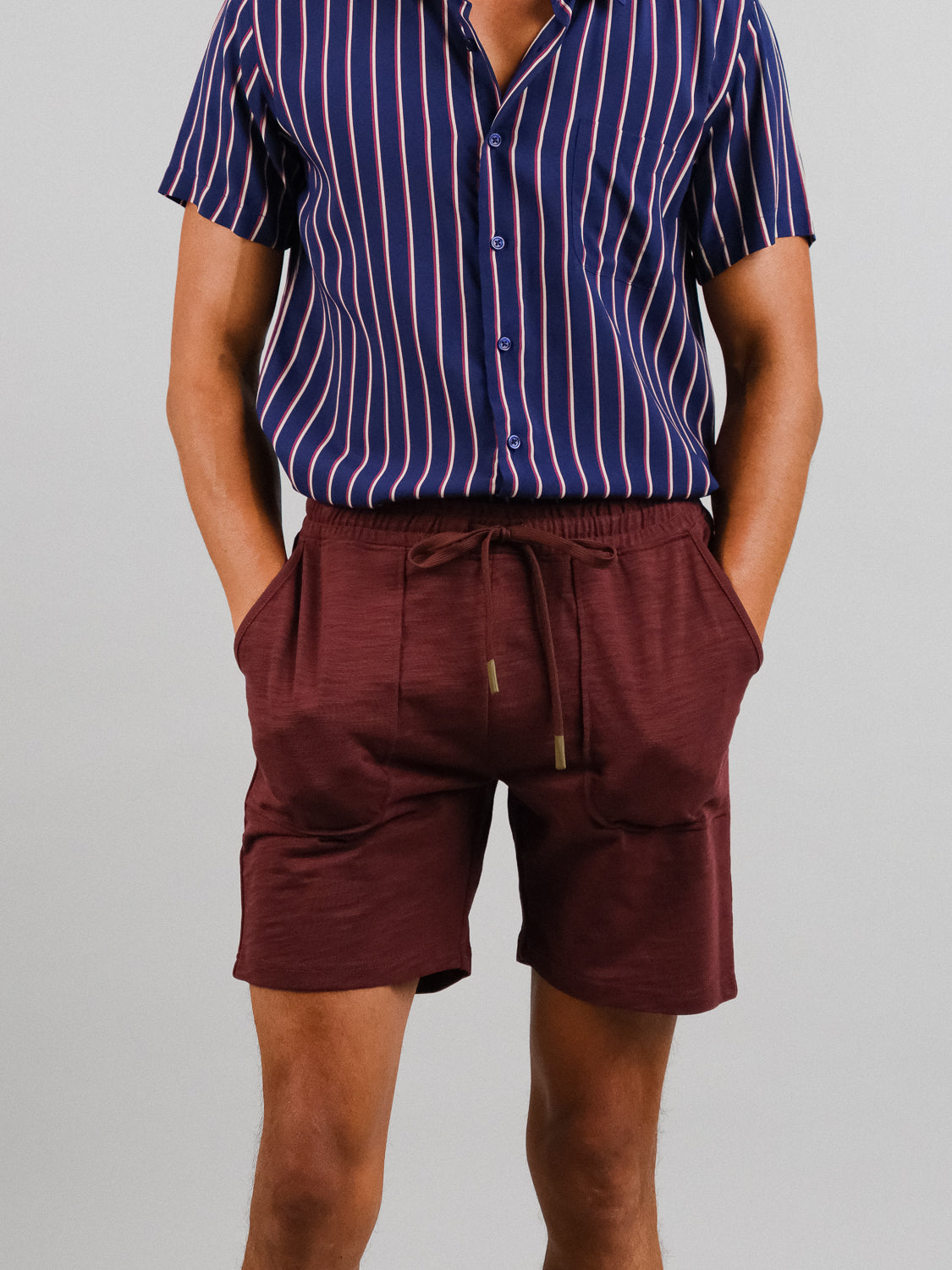 An individual stands against a light gray backdrop, hands in pockets, wearing a blue-and-red striped short-sleeve shirt made of slub cotton and maroon Kahala Slub Cotton Drawstring Shorts by ID Menswear.