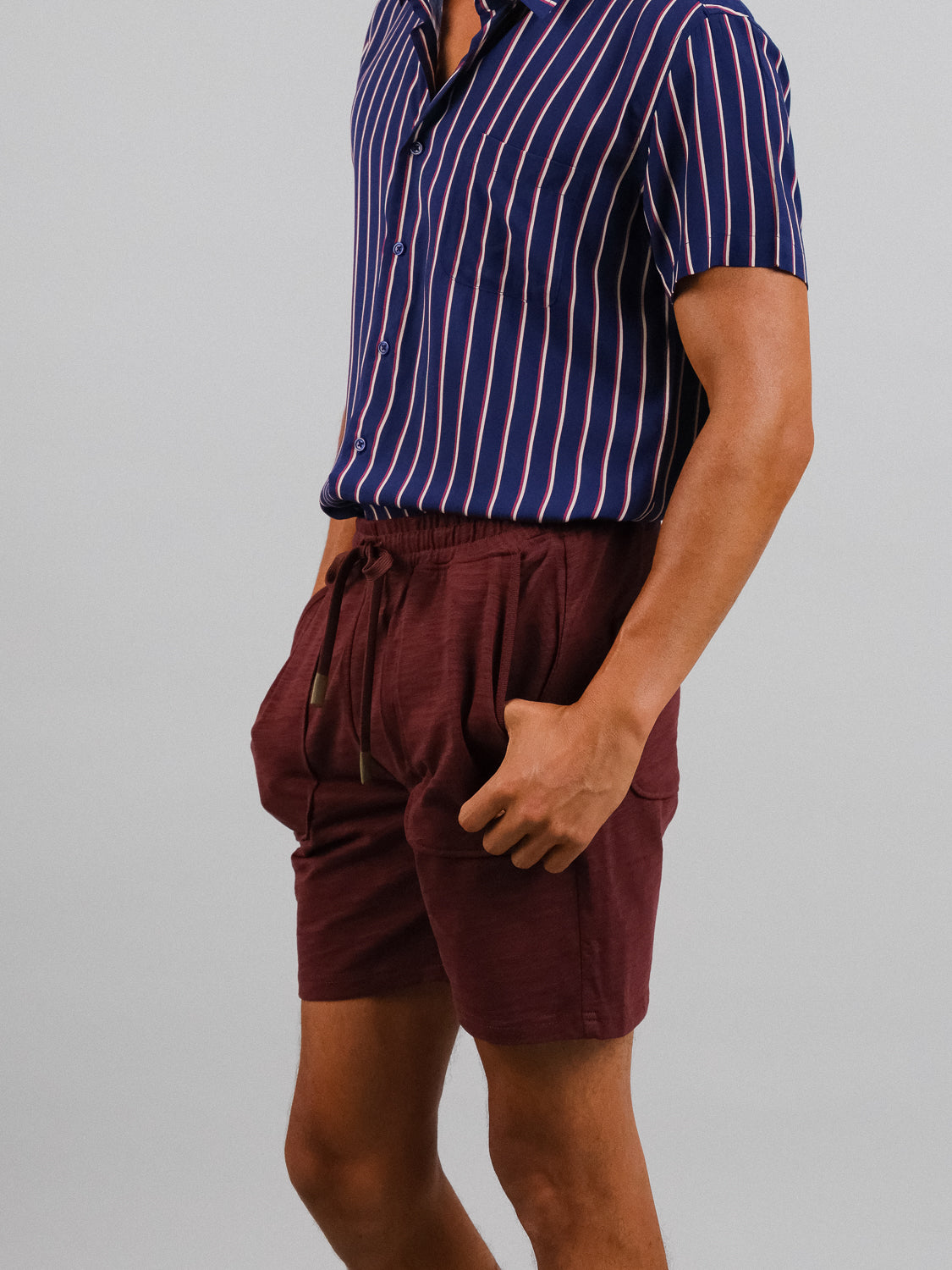 A person in a blue and white striped short-sleeve shirt, made from slub cotton for a relaxed fit, is paired with ID Menswears Kahala Slub Cotton Drawstring Shorts in maroon. They stand against a plain gray background with hands in pockets.