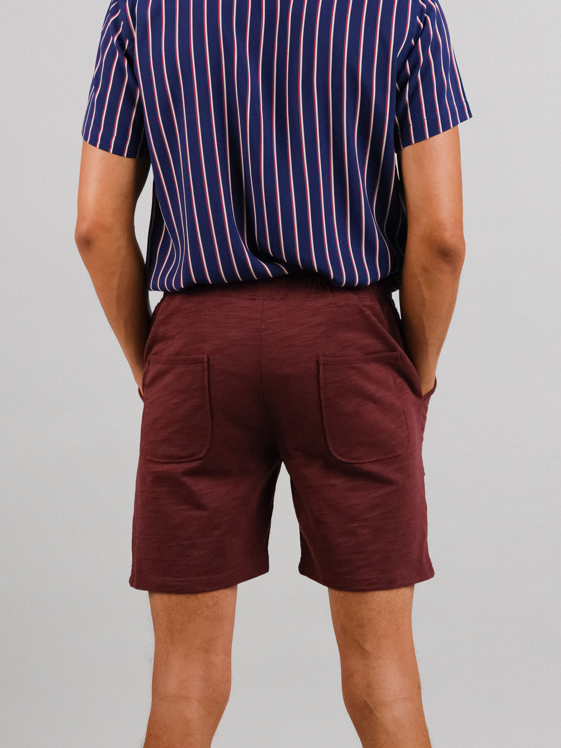 Facing away from the camera, a person wears maroon Kahala Slub Cotton Drawstring Shorts by ID Menswear and a navy shirt with white and red stripes. The shorts have a relaxed fit that complements the casual vibe against the plain light gray background.