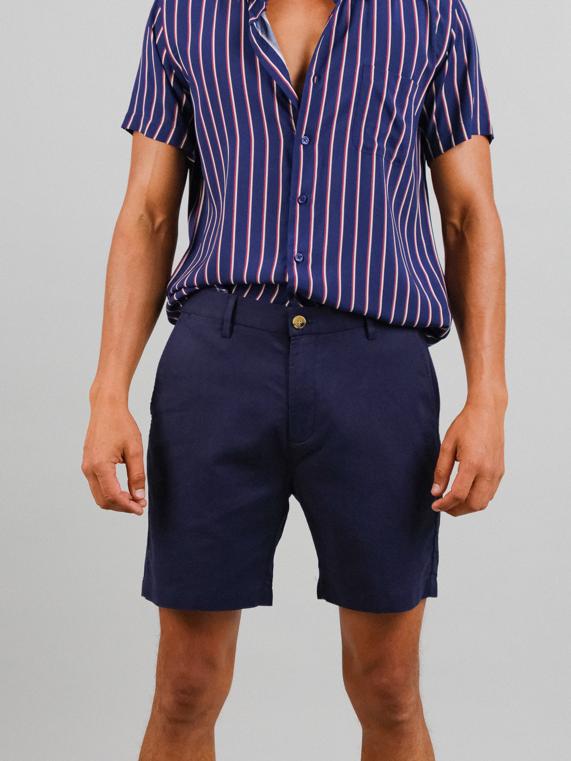 A person in ID Menswears navy blue shirt with red and white stripes and relaxed fit Kerala Linen Blend Chino Shorts exudes a casual, summery vibe. The partially unbuttoned linen-cotton blend shirt reveals a necklace, enhancing the laid-back style.