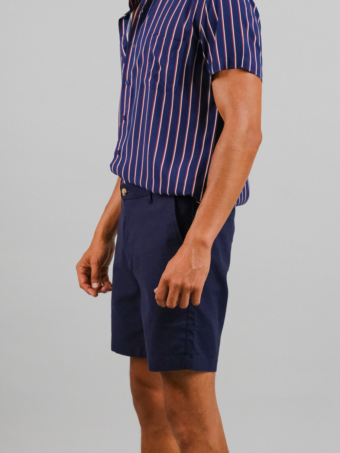 A person wearing a relaxed-fit, navy blue and pink vertically striped short-sleeve shirt pairs it with ID Menswears Kerala Linen Blend Chino Short, standing sideways against a plain gray background.