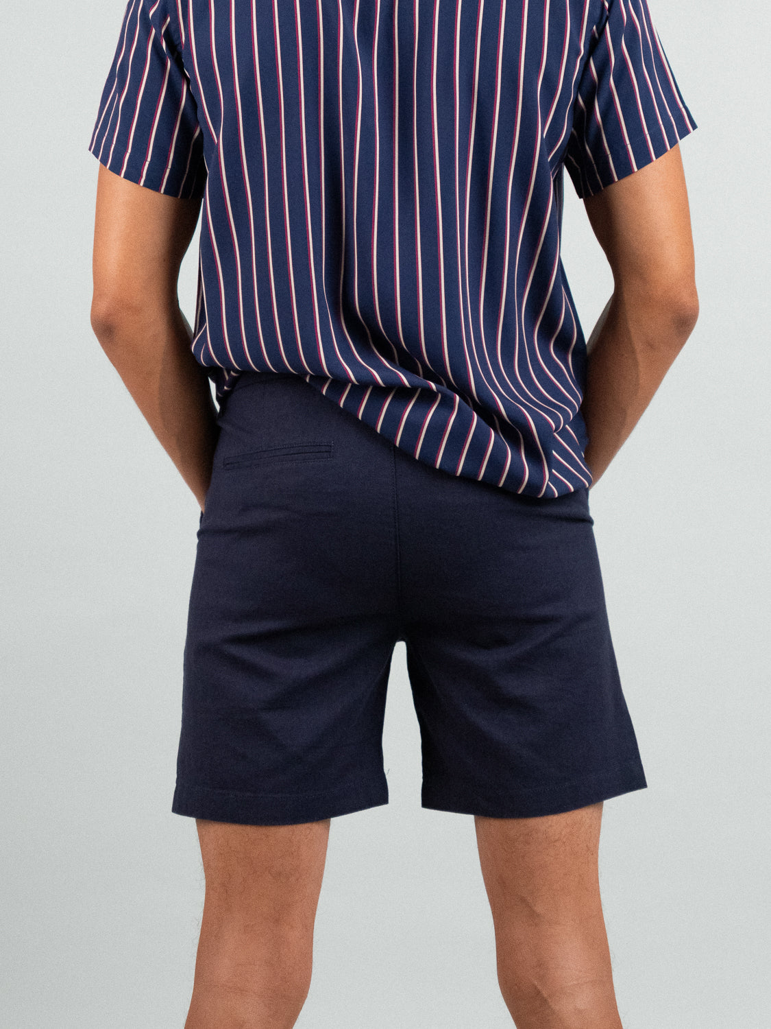 A person is seen from the back wearing a navy and pink striped short-sleeve shirt with ID Menswears Kerala Linen Blend Chino Shorts in dark navy, creating a relaxed fit against a plain gray background.