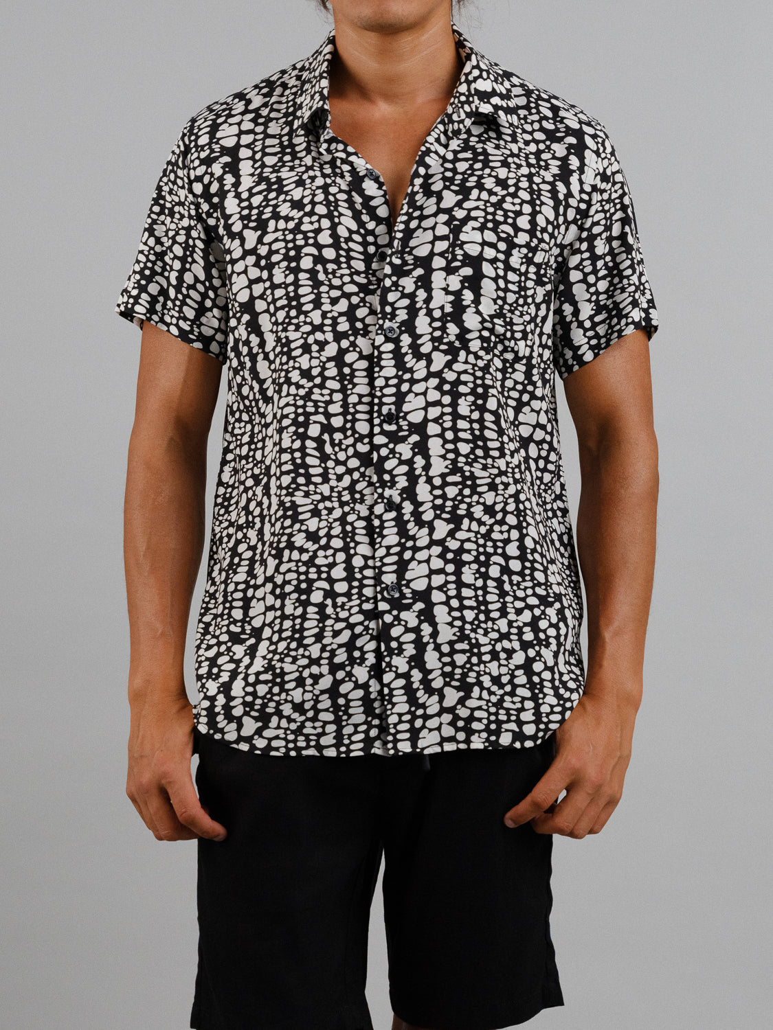 In front of a plain gray background, an individual wears ID Menswears Shimmer Short Sleeve Printed Rayon Shirt with vibrant black and white abstract prints, paired perfectly with black shorts for warm weather.