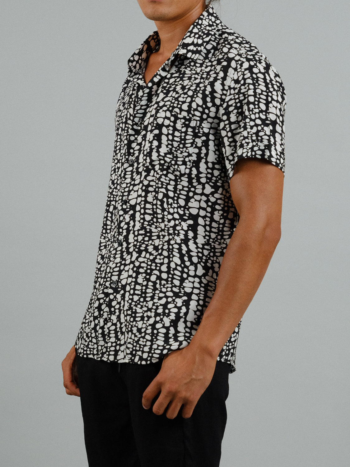 An individual wears ID Menswears Shimmer Short Sleeve Printed Rayon Shirt, showcasing a black-and-white abstract pattern of small irregular white shapes against a plain gray background. The persons face is hidden, highlighting the vibrant design ideal for warm weather.
