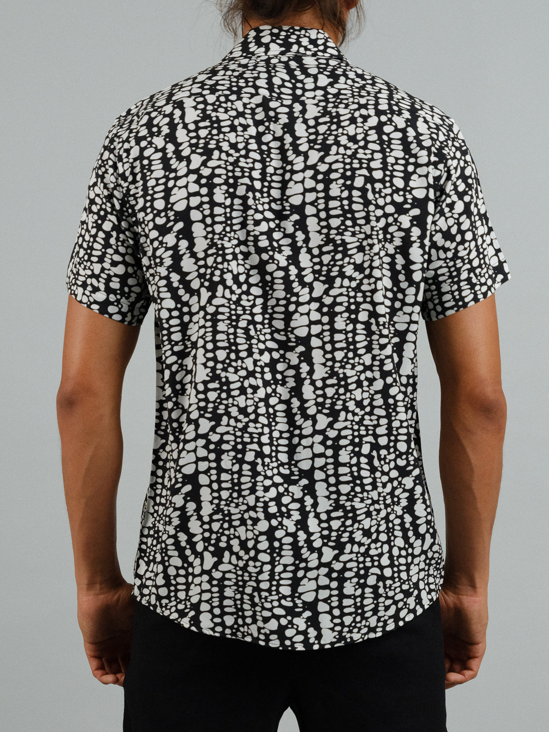 Rear view of someone wearing the Shimmer Short Sleeve Printed Rayon Shirt by ID Menswear. This stylish black and white abstract dot and line pattern is perfect for warm weather. The person stands with relaxed arms against a plain gray background.