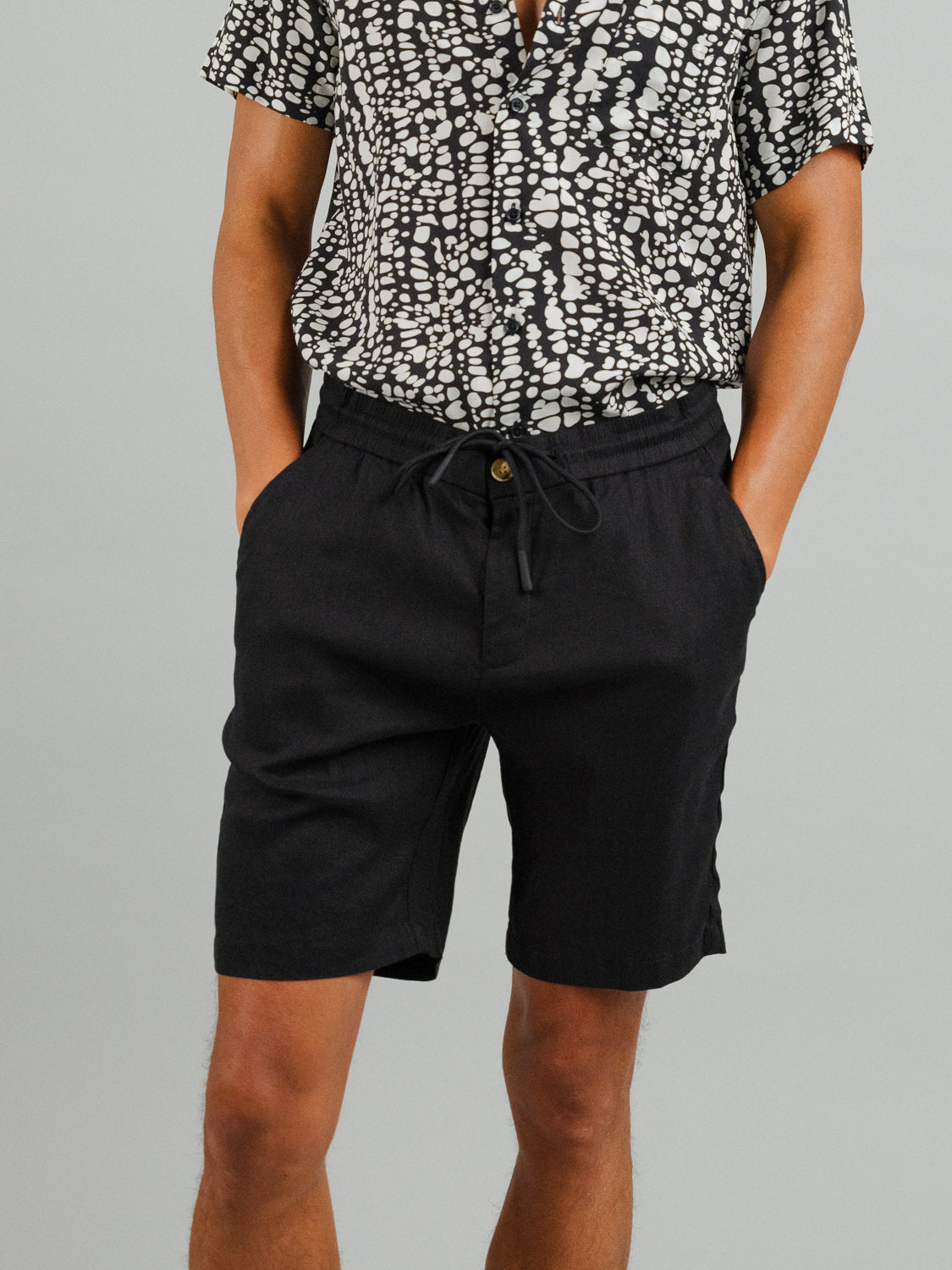 Against a plain backdrop, someone wears a black and white patterned short-sleeve shirt paired with ID Menswears cozy Driggs Linen Blend Shorts—ideal for warm weather. Their hands rest casually in their pockets.