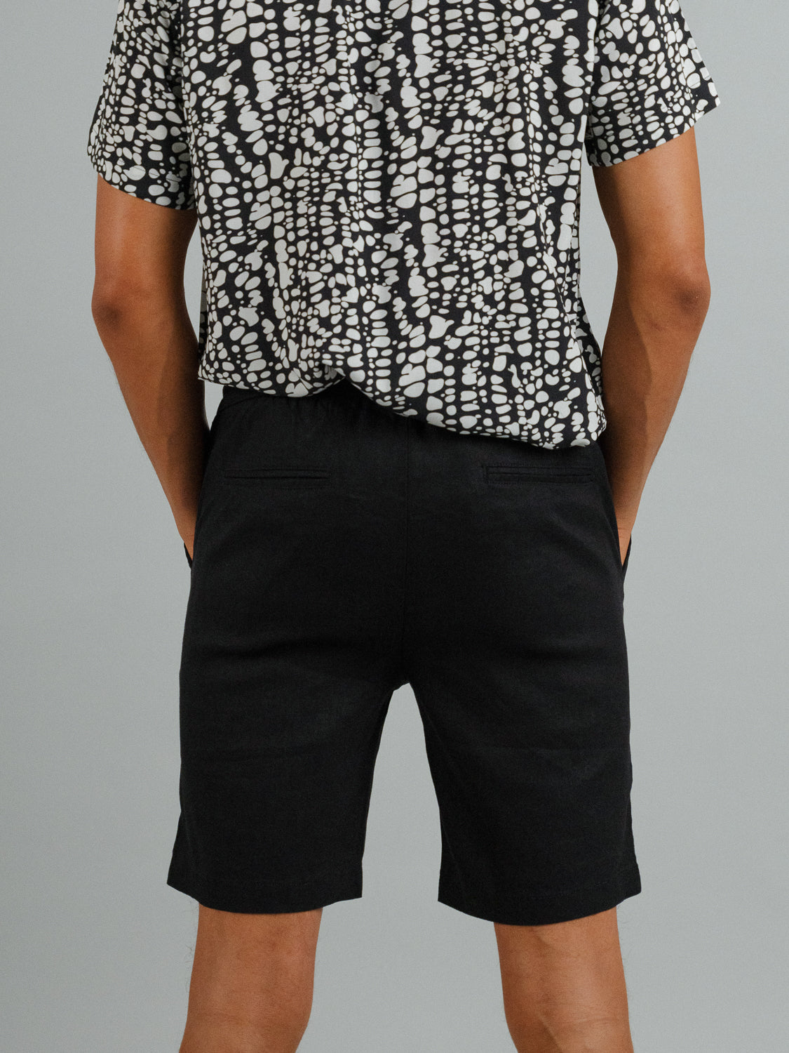 Against a plain gray background, a person stands with hands in pockets, wearing ID Menswears Driggs Linen Blend Shorts and a black and white patterned short-sleeve shirt, ideal for warm weather.
