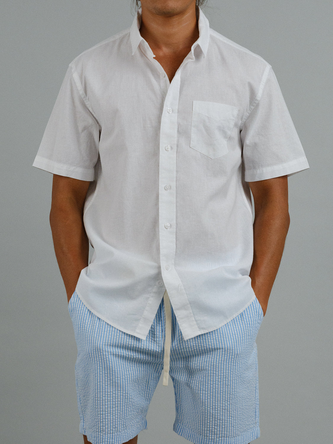 Against a gray background, a person wears the ID Menswear Ravi Linen Cotton Short Sleeve Shirt, featuring a practical front pocket. The crisp white shirt is made from a comfortable linen-cotton blend and paired with light blue striped shorts, hands in pockets.