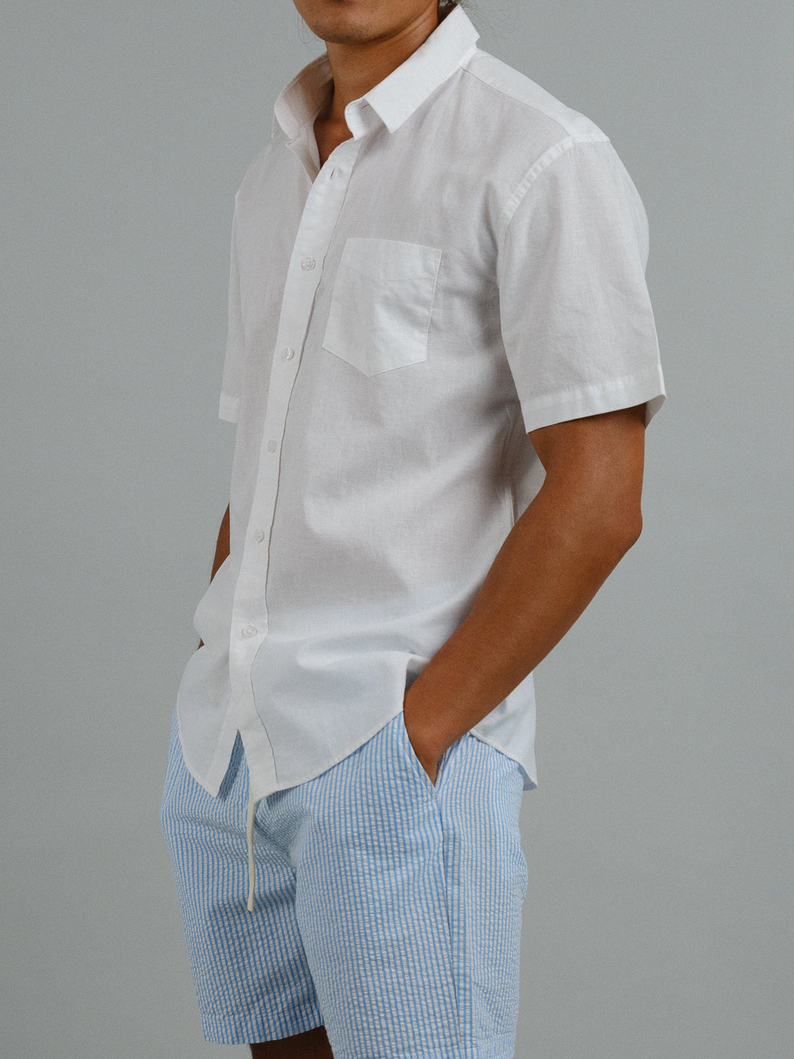 Against a gray backdrop, someone sports ID Menswears Ravi Linen Cotton Short Sleeve Shirt in white, paired with light blue checkered shorts. The linen-cotton blend offers breathable style as they tuck their hands into pockets for a relaxed vibe.