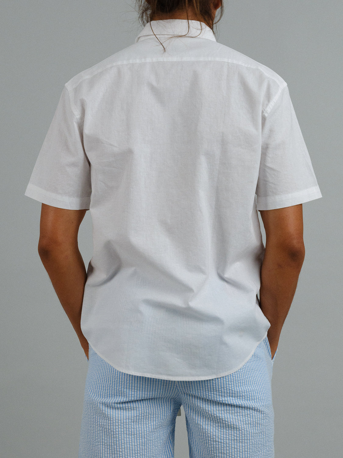 Rear view of a person in an ID Menswear Ravi Linen Cotton Short Sleeve Shirt, paired with light blue striped shorts against a plain gray background.