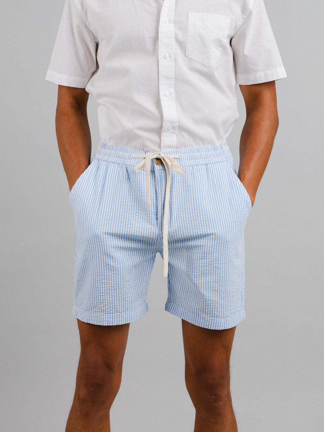 A person wearing a white short-sleeve button-up shirt and ID Menswears Ibiza Seersucker Drawstring Shorts, hands in pockets, ideal for warm weather. The backdrop is plain gray.