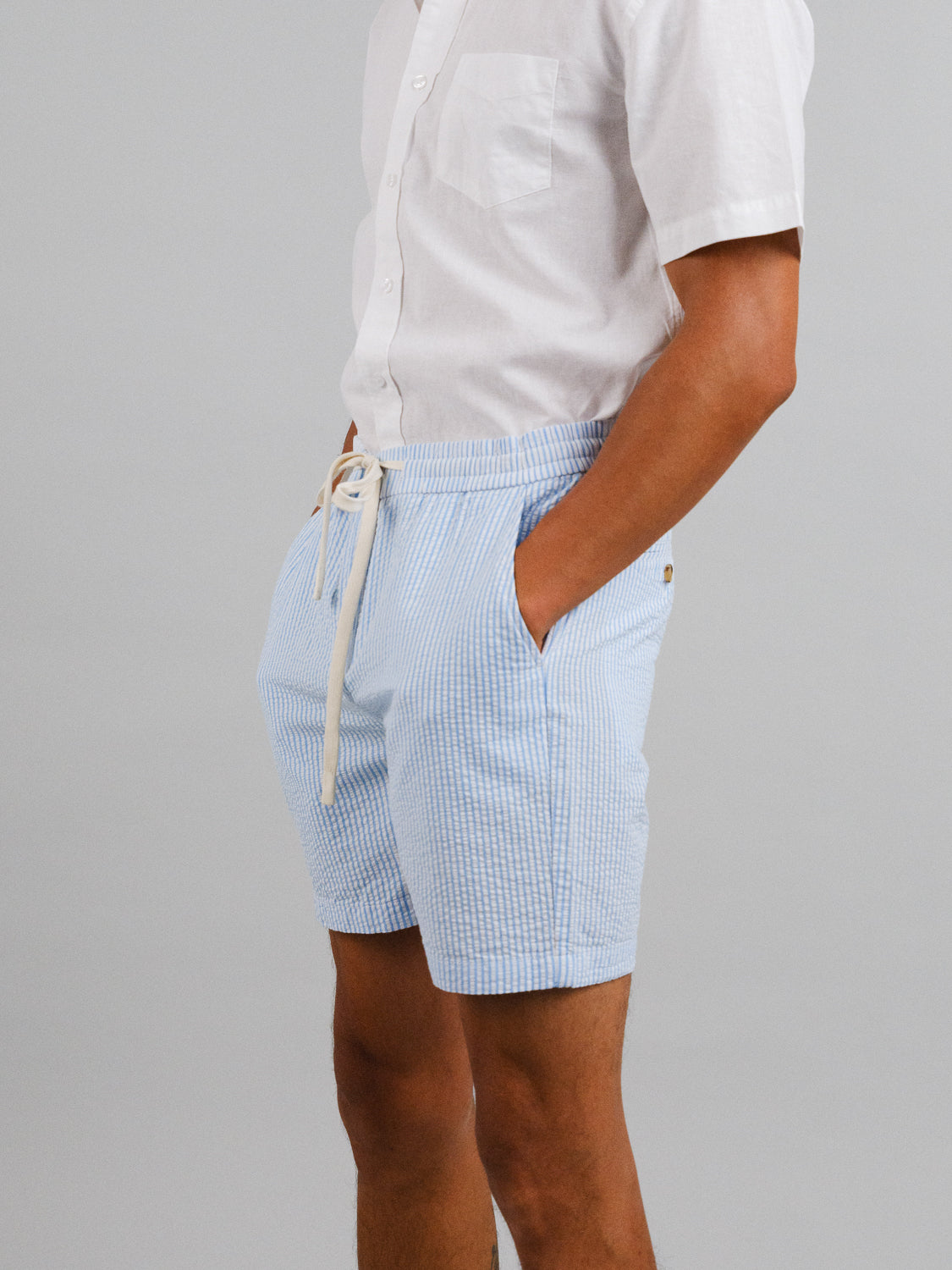 A man stands against a gray background, hands in the pockets of his light blue striped Ibiza Seersucker Drawstring Shorts by ID Menswear. The white short-sleeved shirt perfectly completes his warm-weather wardrobe look.