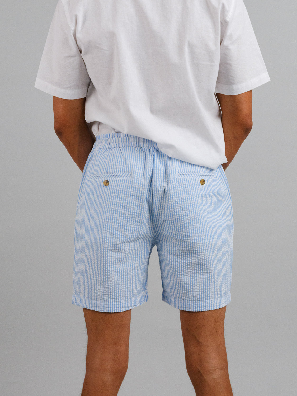 A person stands with their back to a plain gray wall, wearing a white T-shirt and ID Menswears blue-and-white striped Ibiza Seersucker Drawstring Shorts, featuring an elastic waistband and two back pockets with buttons—ideal for warm weather.
