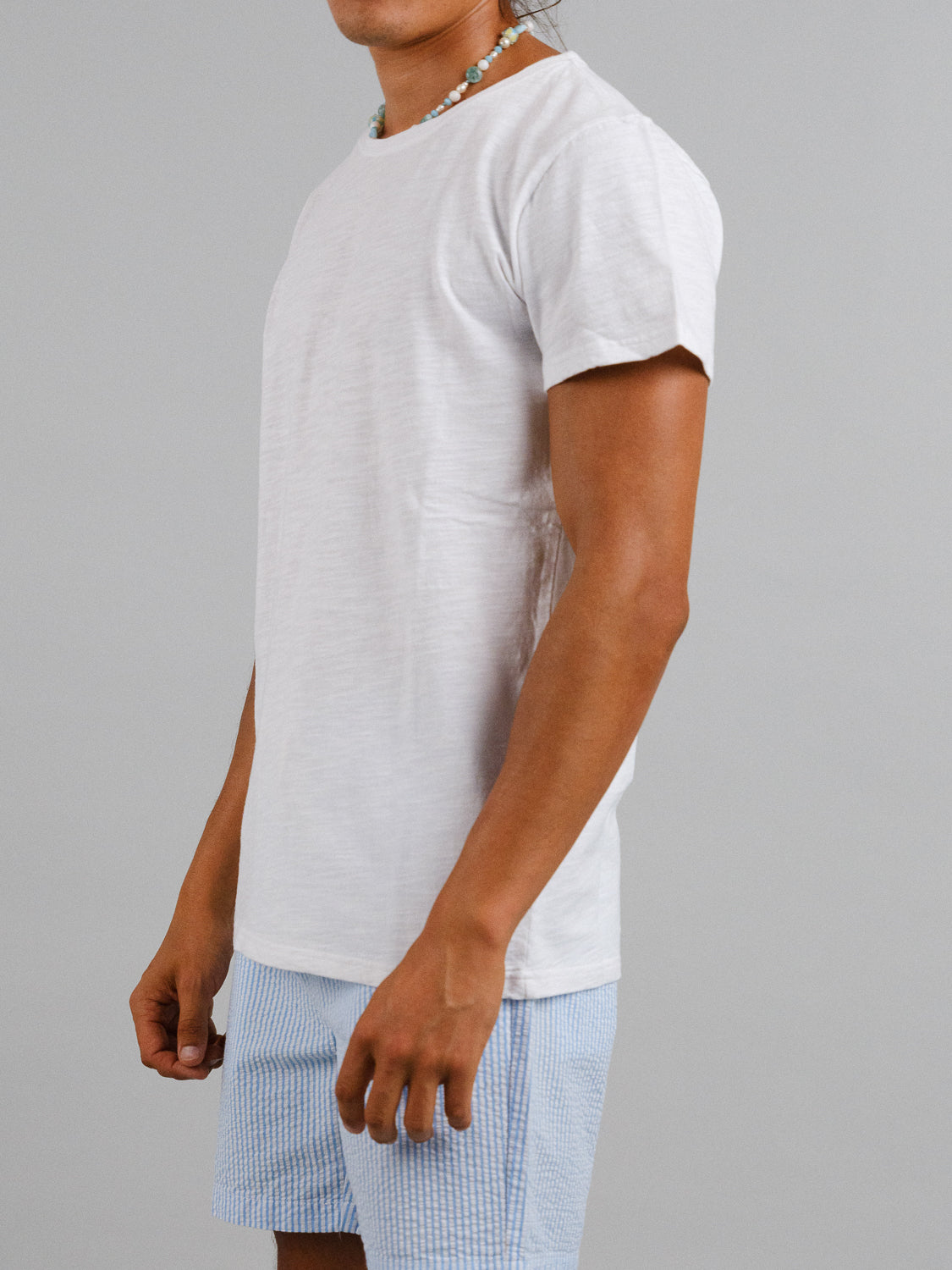 An individual in an everyday outfit, wearing a Mini Flip Slub Yarn Tee by ID Menswear, light blue checkered shorts, and a beaded necklace stands against a plain gray backdrop. Slightly sideways with relaxed arms, they showcase a casual vibe.