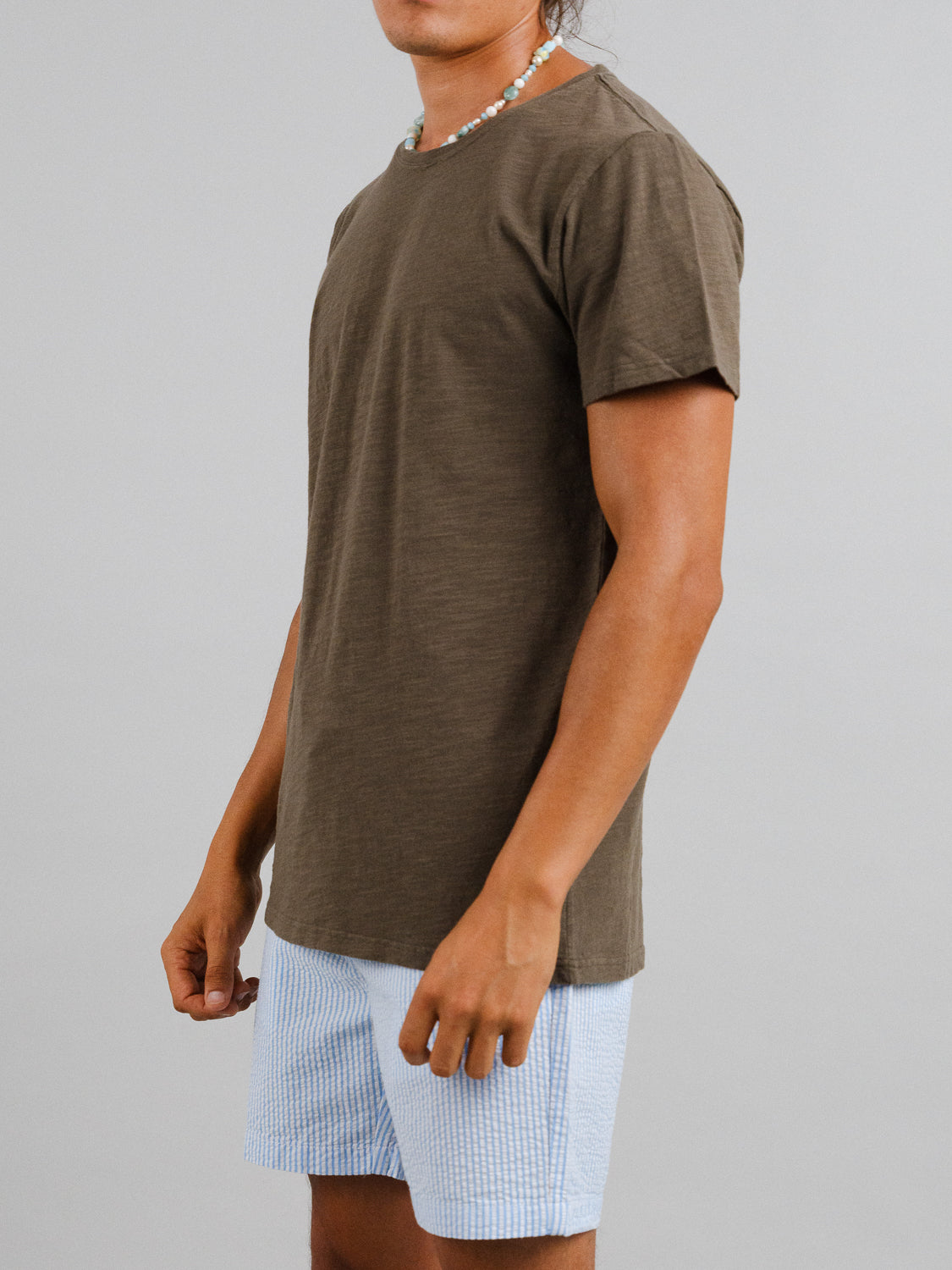 A person in a casual style wears ID Menswears Mini Flip Slub Yarn Tee in brown, along with light blue shorts and a necklace. They pose slightly angled to the side against a plain white backdrop.