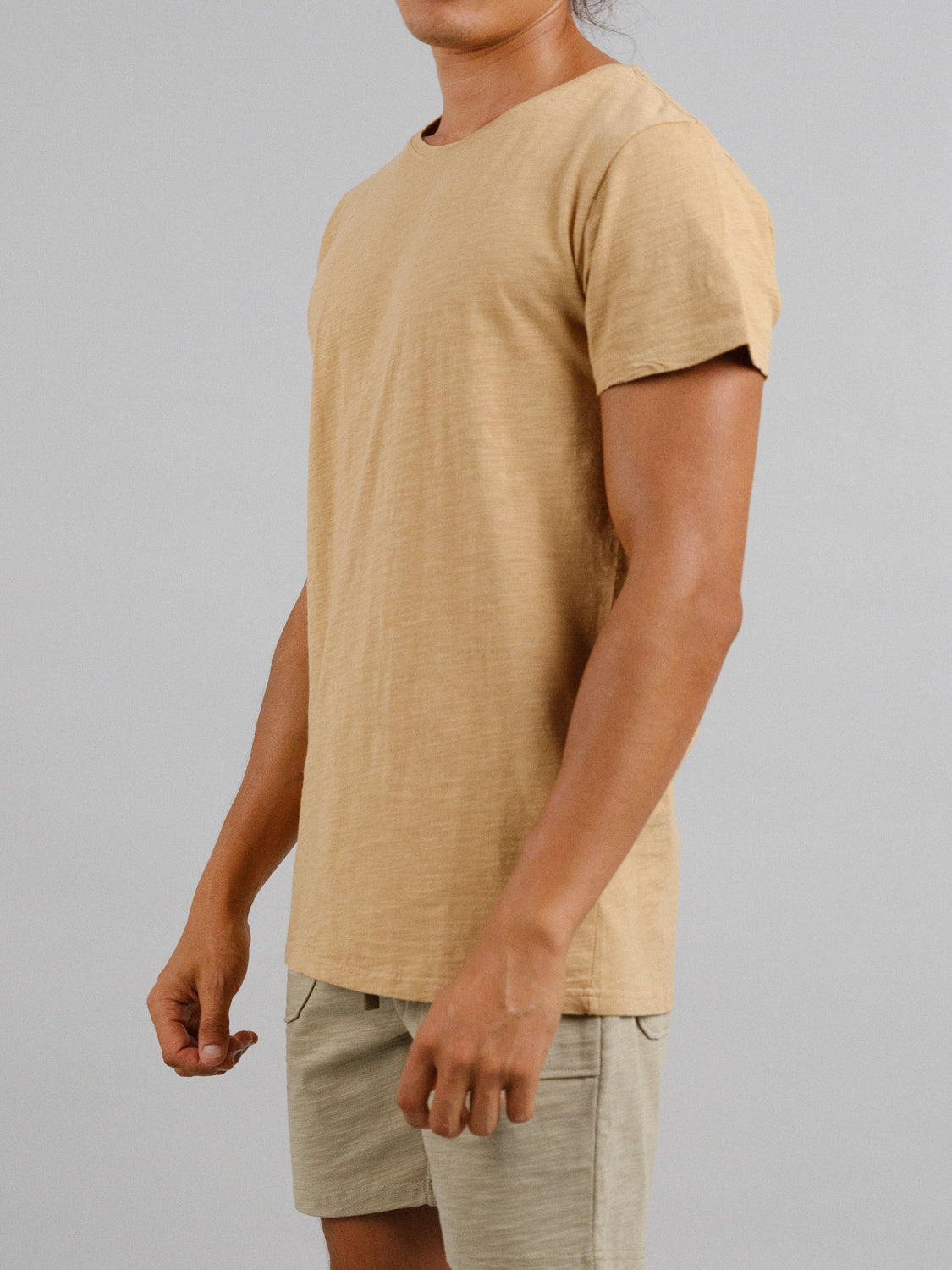 Dressed in a beige Mini Flip Slub Yarn Tee from ID Menswear and light khaki shorts, the person projects a relaxed vibe with arms gently bent against a simple gray backdrop.