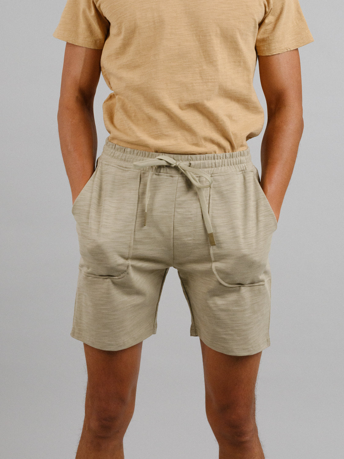 A person in a light brown slub cotton t-shirt and ID Menswears Kahala Slub Cotton Drawstring Short in relaxed fit beige stands with hands in pockets against a plain gray background.