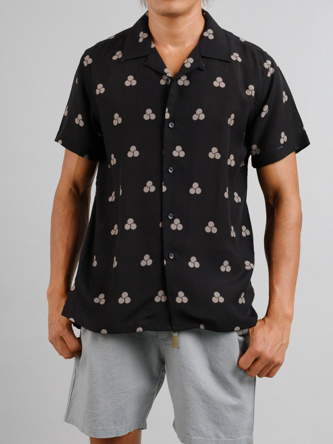 Someone is wearing the ID Menswear Mae Camp Collar Printed Shirt, a black short-sleeve shirt with beige geometric designs made from a breathable Tencel-linen mix, paired perfectly with light gray shorts against a plain background.