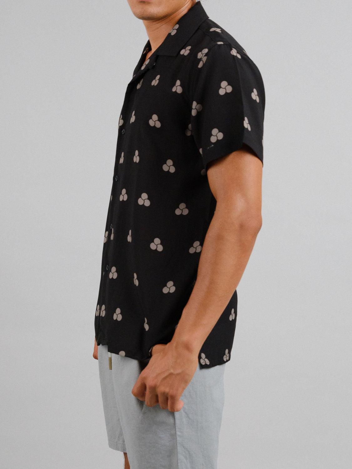 A person models an ID Menswear Mae Camp Collar Printed Shirt in a tencel-linen blend with a beige abstract pattern on black. Their face isnt shown, and the breathable shirt pairs effortlessly with light gray shorts against a plain, neutral backdrop.
