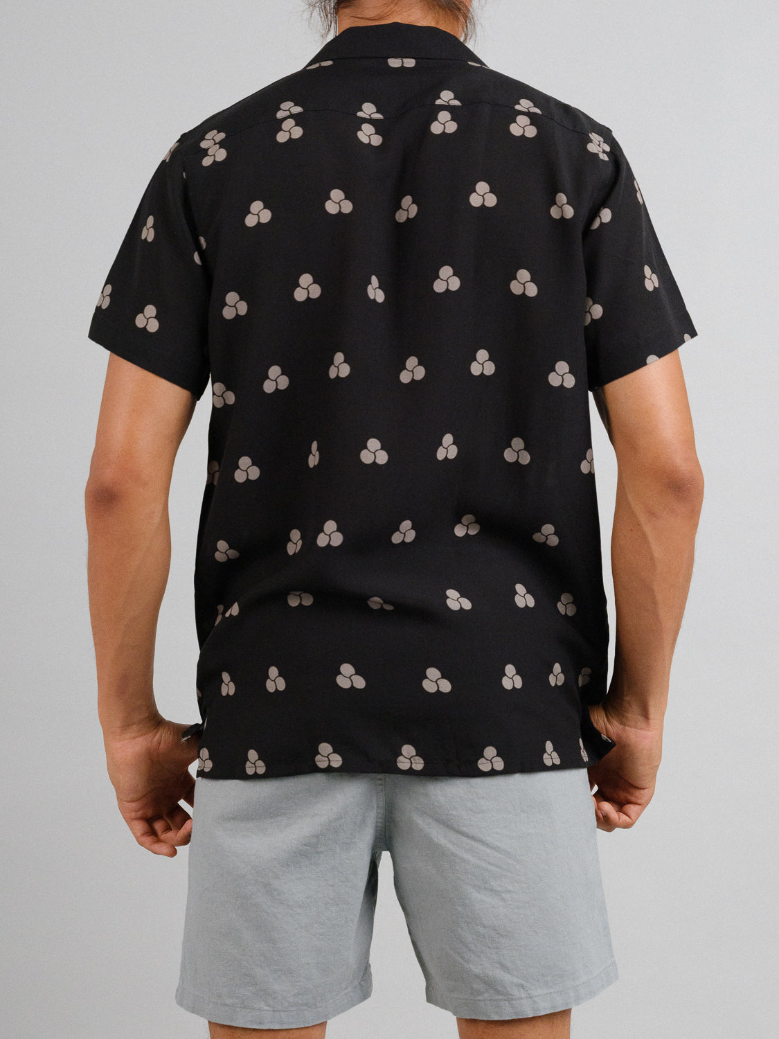 A person stands against a plain gray backdrop, wearing the Mae Camp Collar Printed Shirt by ID Menswear. The shirt features black with light gray atomic symbols paired with breathable light gray tencel-linen shorts. Theyre facing away from the viewer.