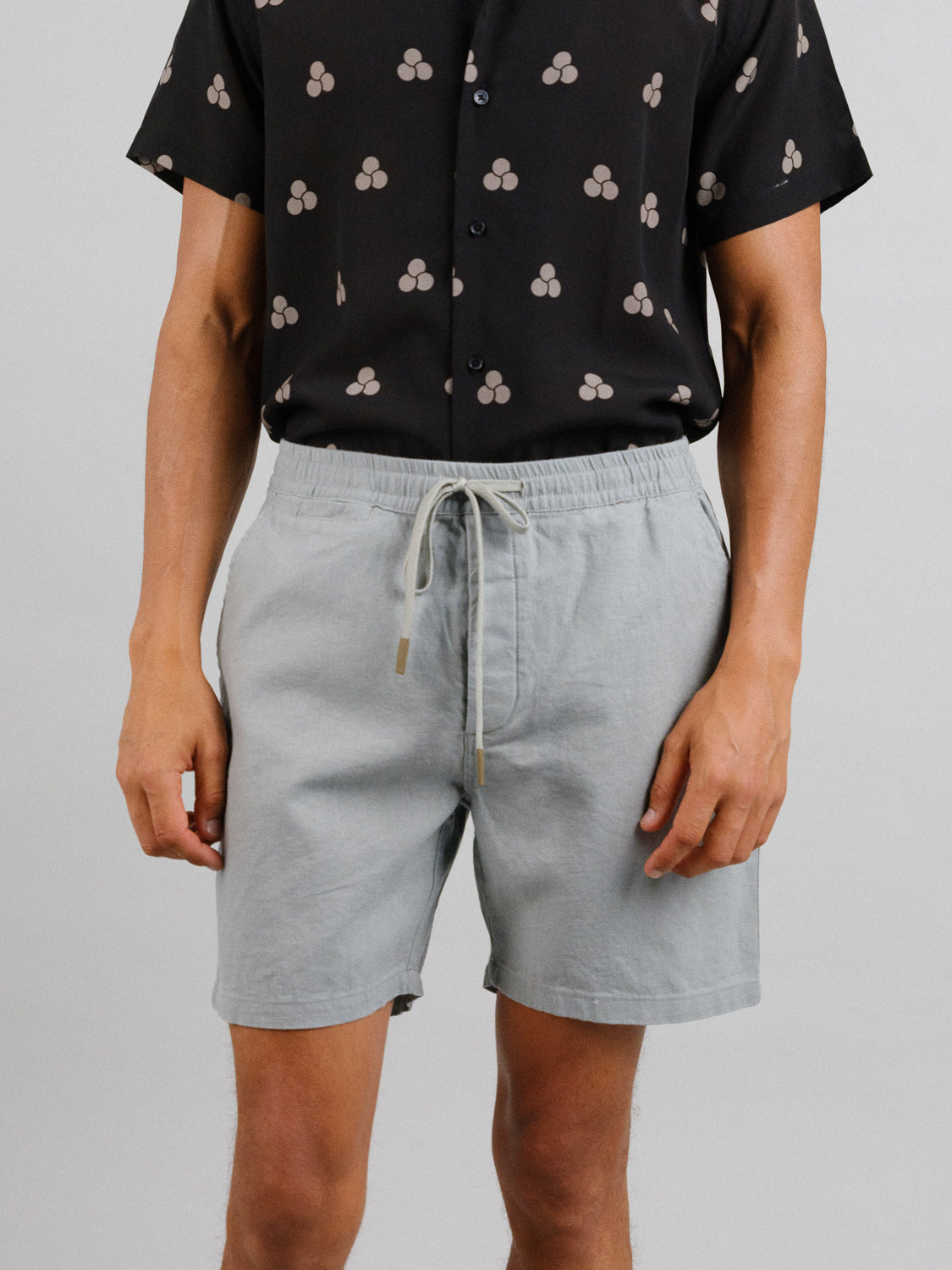 A person dressed in a black short-sleeved shirt with beige circle patterns and ID Menswears Kotao Linen Blend Drawstring Short in light gray stands against a plain background, highlighting their torso and legs.
