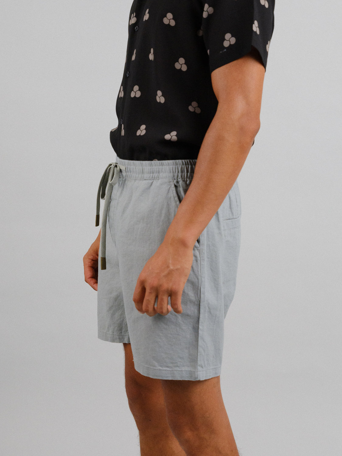 A person stands against a plain background wearing ID Menswears Kotao Linen Blend Drawstring Shorts in light gray and a black shirt adorned with light symbols.