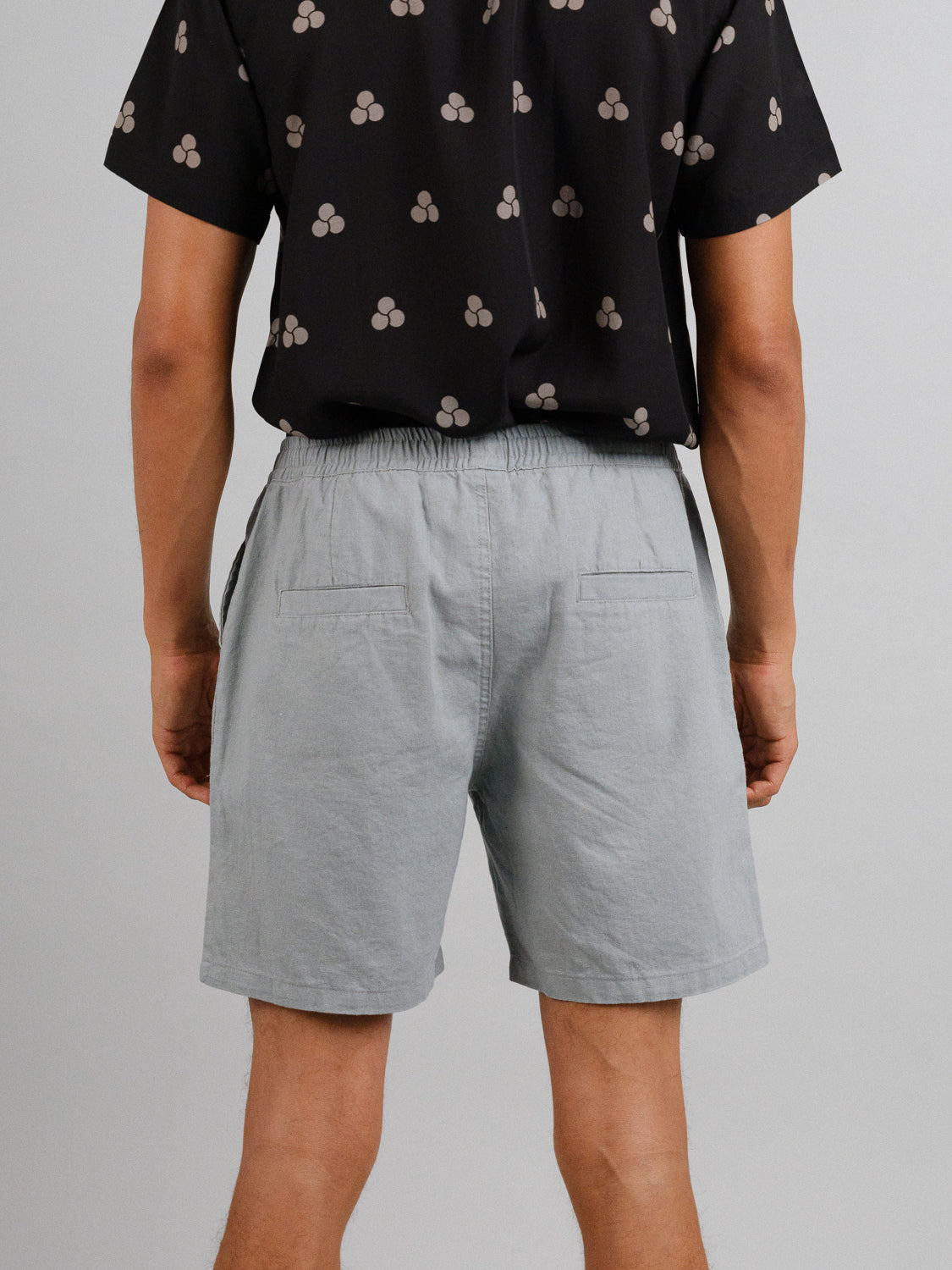 Seen from the back, a person wears a black shirt with a white pattern and Kotao Linen Blend Drawstring Shorts in light gray by ID Menswear. The outfit is perfect for relaxed summer days against a plain background.