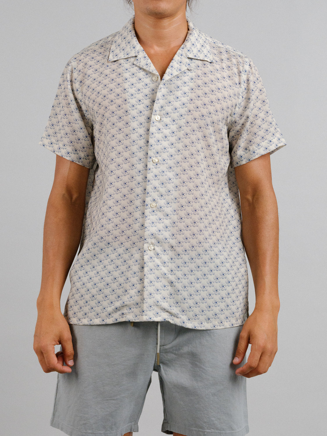 A person wears the Sasa Camp Collar Printed Shirt by ID Menswear, a breathable, light-colored Tencel-linen blend with a relaxed fit and subtle pattern, paired with light gray shorts against a plain gray background.