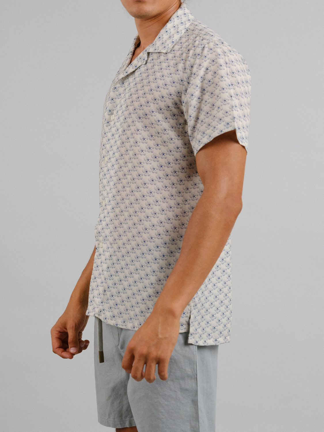 A person in the Sasa Camp Collar Printed Shirt by ID Menswear and light gray shorts stands relaxed against a plain background. The breathable Tencel-linen blend fabric complements their laid-back posture, though their head is out of frame.