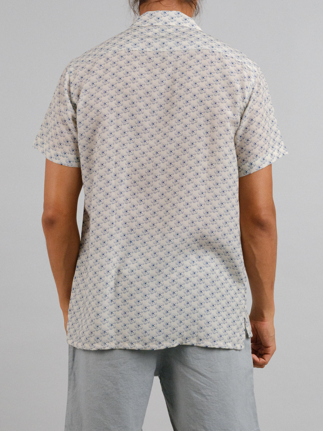 A person is seen from behind wearing the Sasa Camp Collar Printed Shirt from ID Menswear; its a short-sleeved, cream-colored shirt with a small blue pattern, paired with light gray pants. The tencel-linen blend provides breathable comfort against the plain gray background.