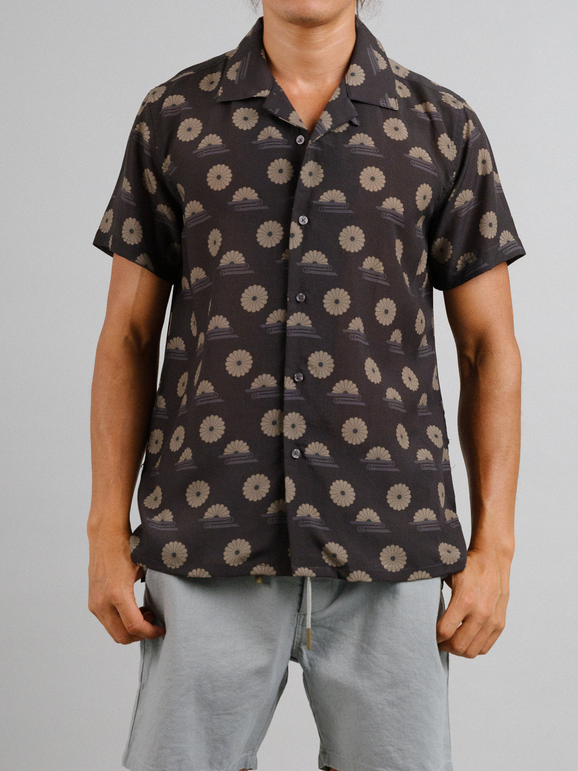 A man wears the ID Menswear Kiku Camp Collar Printed Shirt, a short-sleeved, button-up shirt with a dark floral print, ideal for summer. Made from a Tencel linen blend, it pairs well with light gray shorts against a plain background. His head is not visible in the image.