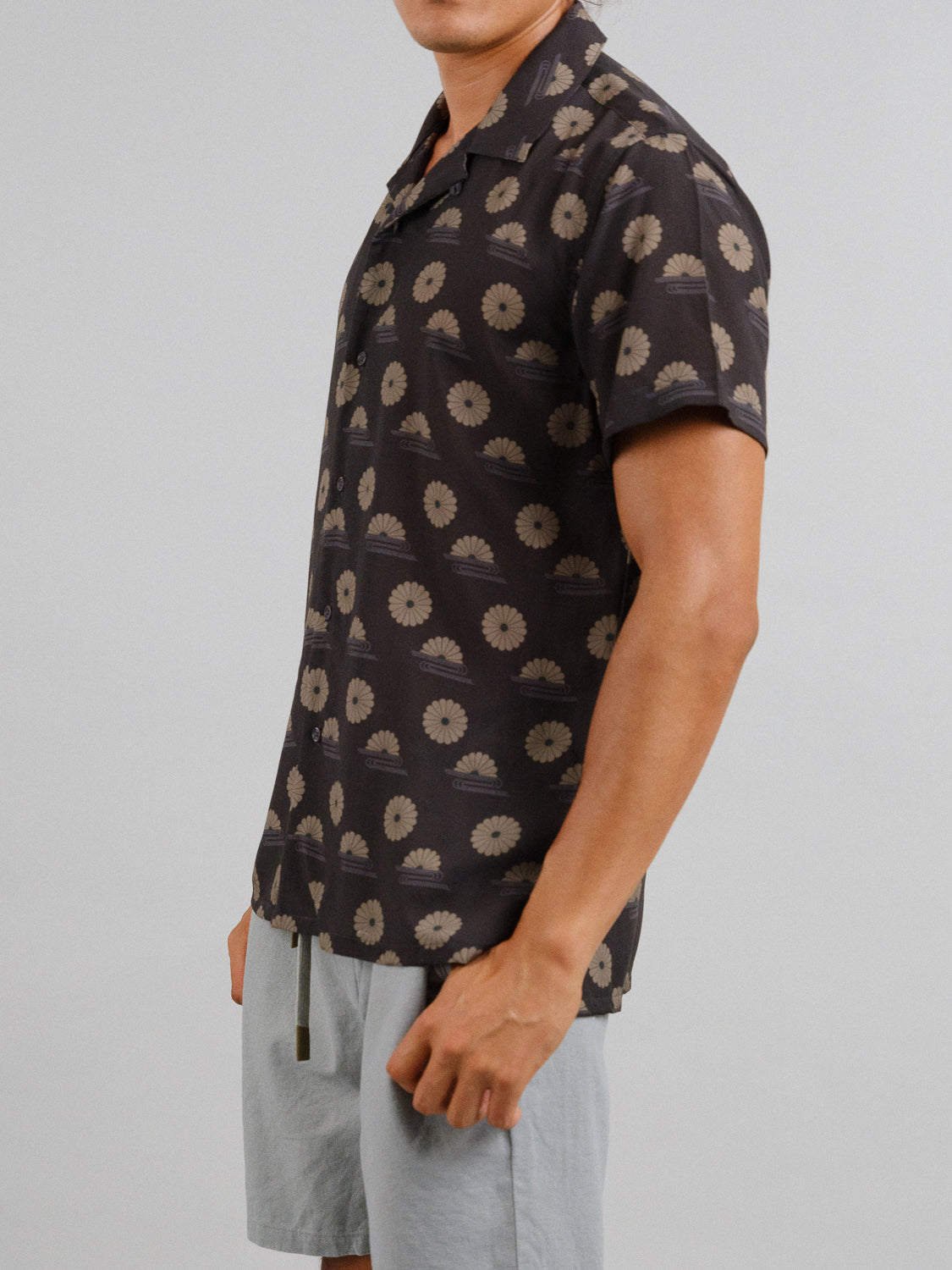 A person wears the Kiku Camp Collar Printed Shirt by ID Menswear, featuring a dark backdrop with beige floral designs, paired with light-colored shorts. Ideal for warm weather outings, the short-sleeved summer shirt is showcased against a plain background.