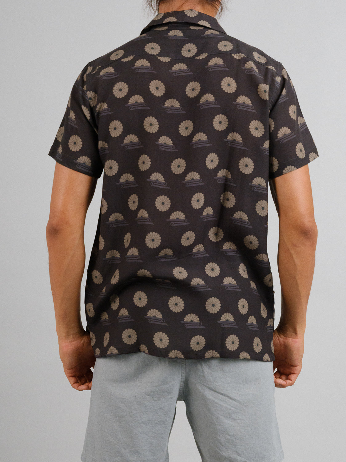 A person faces away from the camera, wearing the Kiku Camp Collar Printed Shirt by ID Menswear—a short-sleeve shirt with a tan floral pattern on a dark Tencel linen blend. Light gray shorts complement the look against a plain gray wall.