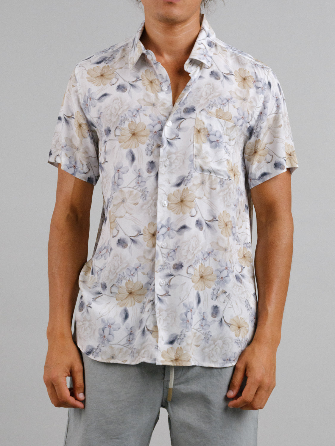 A person wearing the Tobe Short Sleeve Printed Rayon Shirt by ID Menswear in beige and gray, featuring a subtle floral print with a left-side pocket, paired with gray pants. The top button is undone against the plain gray backdrop, highlighting its warm-weather vibe.