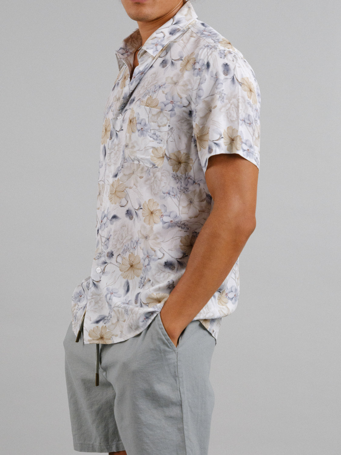 A man stands against a plain background wearing the Tobe Short Sleeve Printed Rayon Shirt by ID Menswear, featuring vibrant floral prints. Paired with gray shorts, he casually rests his right hand in his pocket, embodying warm weather style.