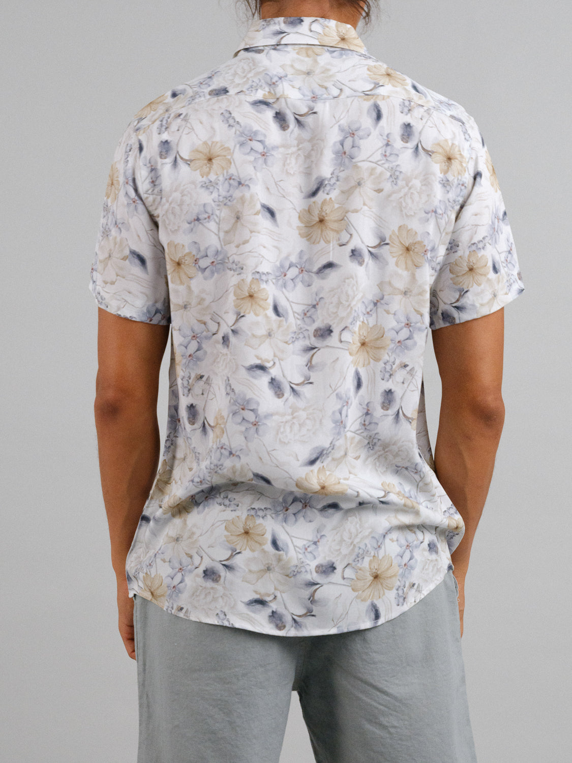 With their back to the camera, a person with long hair wears a Tobe Short Sleeve Printed Rayon Shirt by ID Menswear, showcasing floral patterns in soft white, beige, and gray tones. Paired with gray pants against a plain background, the outfit exudes a warm weather vibe.