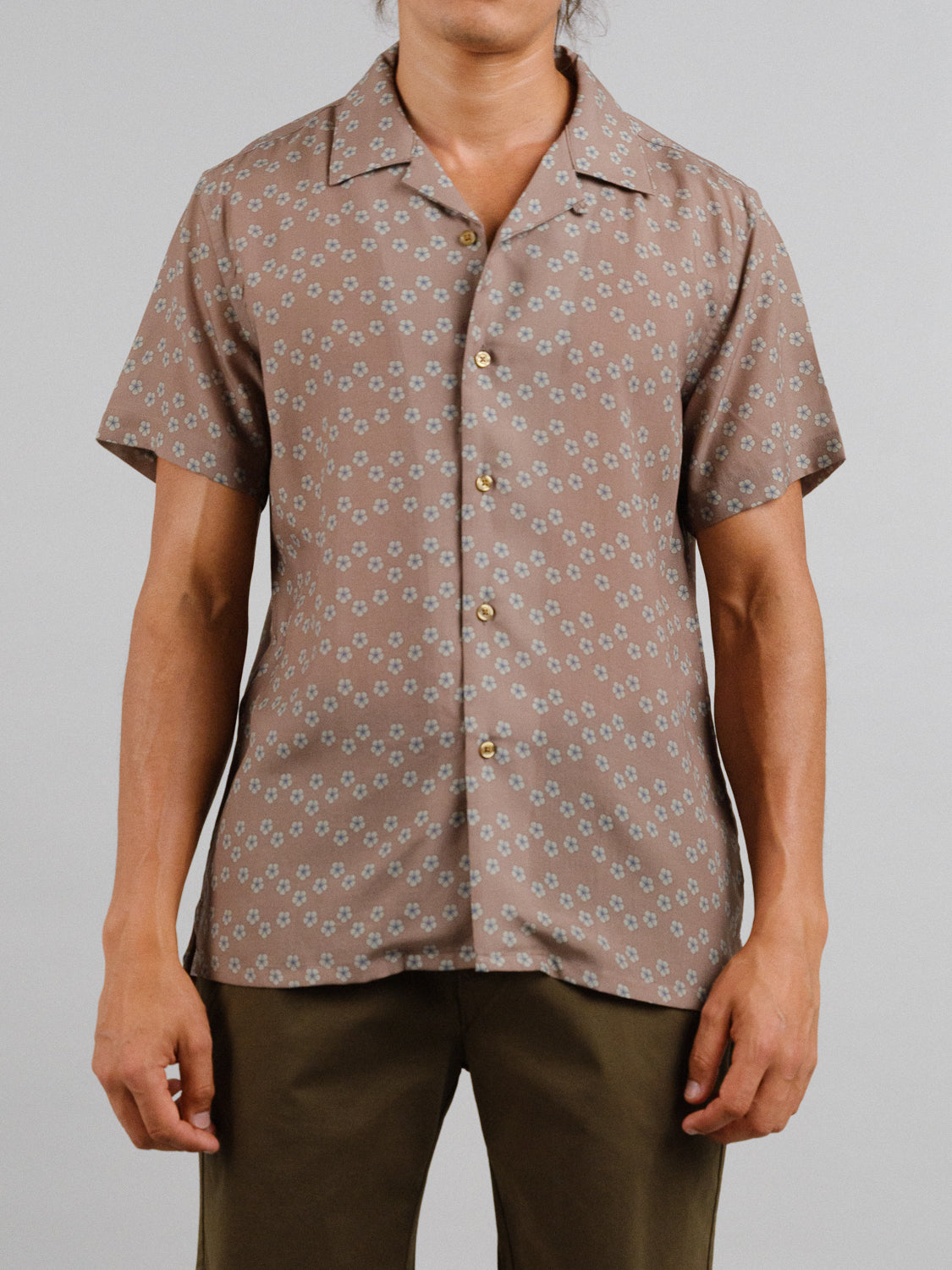 Against a plain backdrop, an individual wears the brown Sai Camp Collar Printed Shirt by ID Menswear, featuring a breathable tencel-linen blend with small white flowers and gold buttons, paired with matching brown pants.