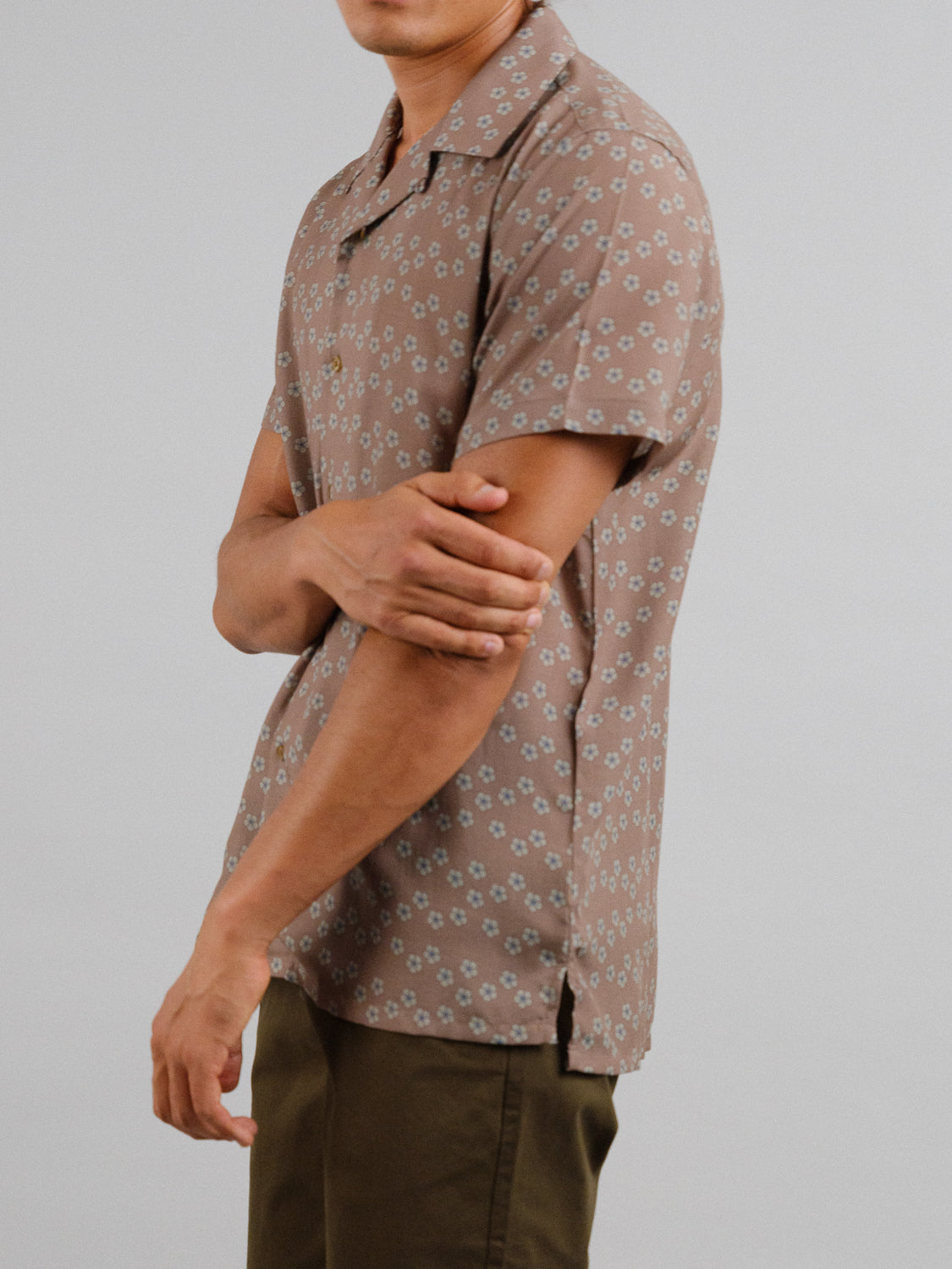 Against a plain backdrop, a person wears the ID Menswear Sai Camp Collar Printed Shirt featuring a small floral pattern and short sleeves. They cross their arms in olive green tencel-linen blend pants, showcasing breathable comfort.