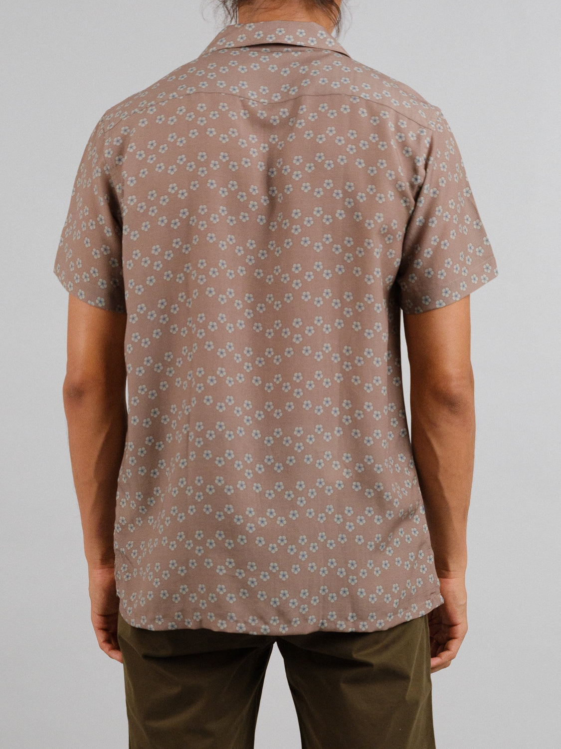 A person with long hair stands facing away, wearing the ID Menswear Sai Camp Collar Printed Shirt, which features a small white floral pattern on brown tencel-linen fabric for breathable comfort, paired with dark green pants against a plain light gray background.